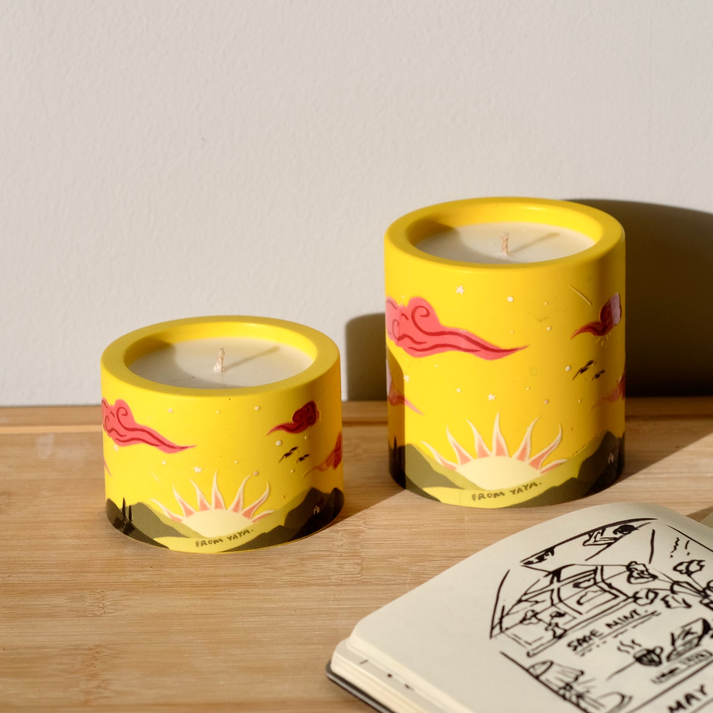 Small and large sunrise essential oil candles side by side on a table, yellow illustration of sun and green mountains.