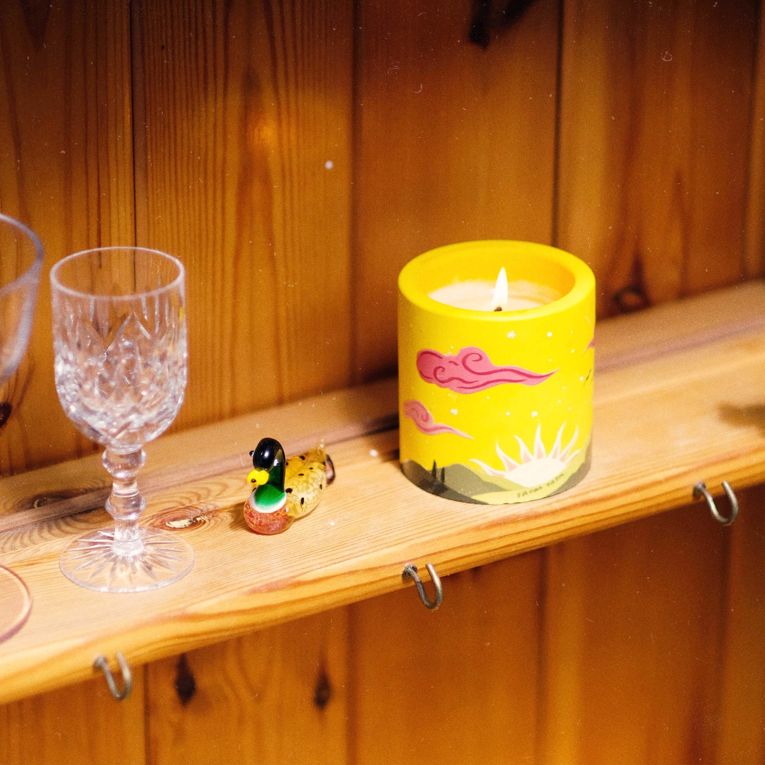 A yellow cylindrical candle with pink and white cloud-like illustrations sits on a wooden shelf next to a crystal wine glass and a small duck figurine, against a wood-paneled background.