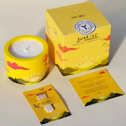 Small yellow natural essential oil candle with illustration of a sunrise over mountains, packaging, matches, and card on a white background.