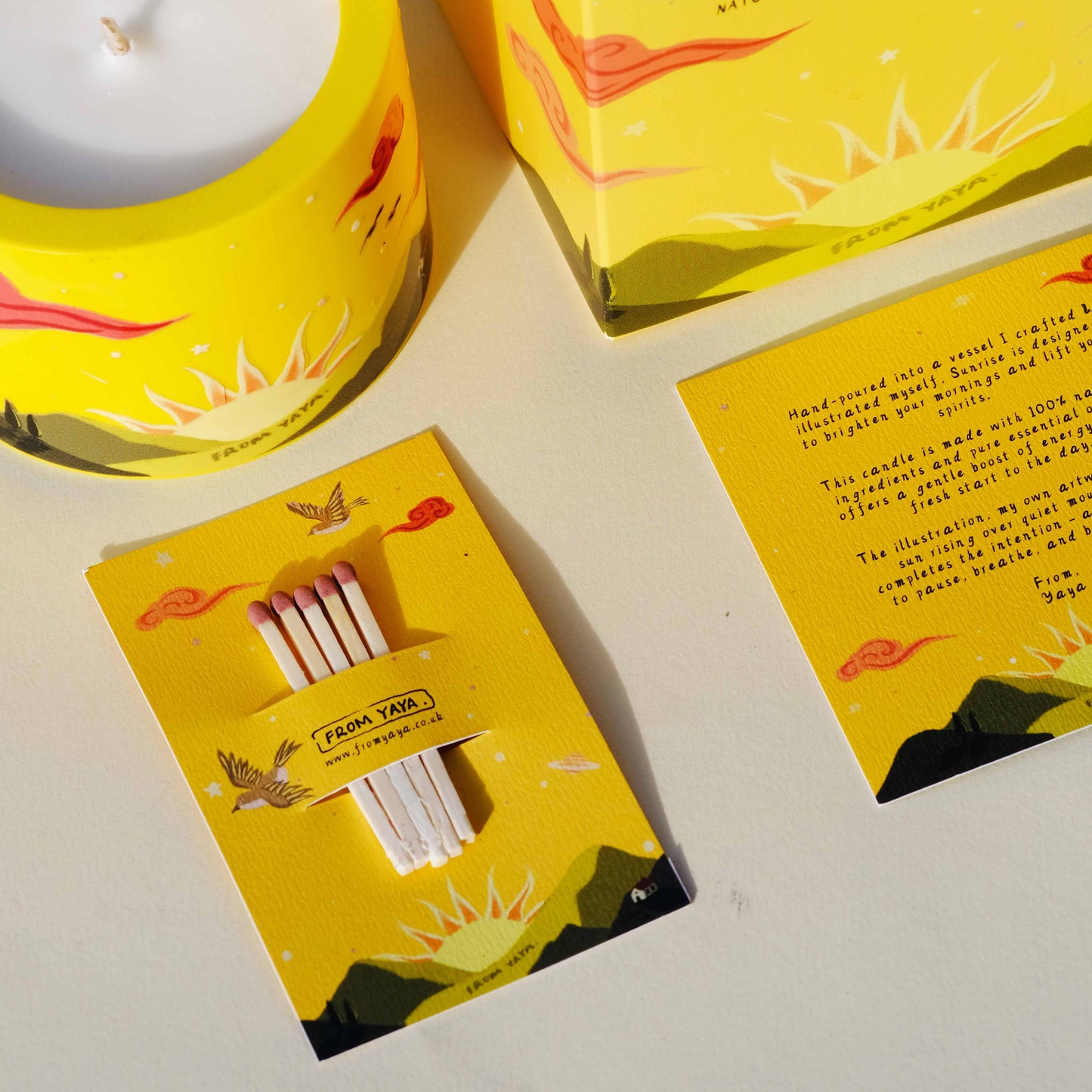 Yellow essential oil candle packaging with matches and scenic illustrations of a sunrise over mountains on a white background.