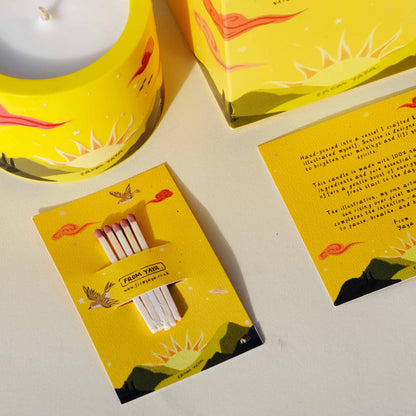 Yellow essential oil candle packaging with matches and scenic illustrations of a sunrise over mountains on a white background.